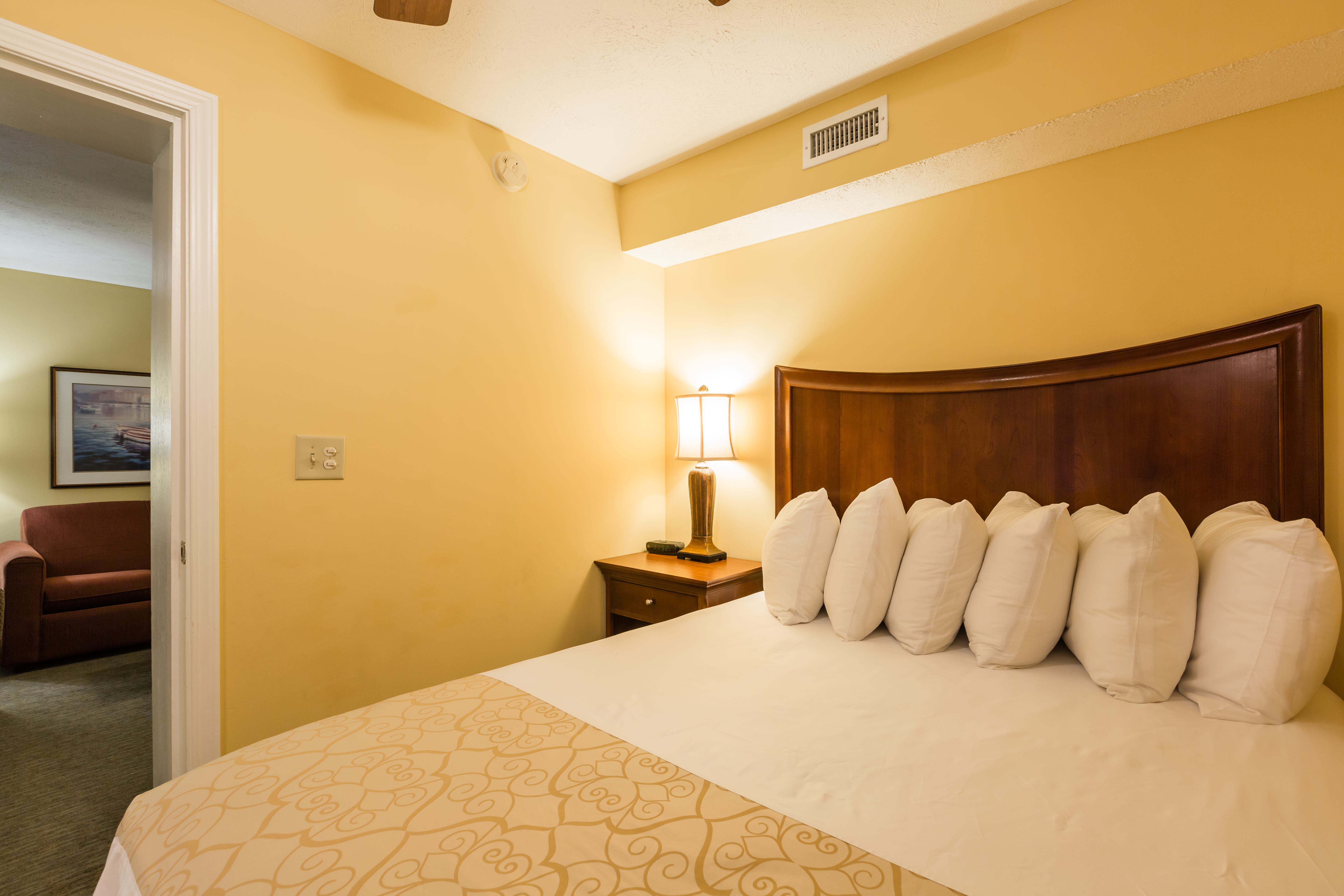 A hotel room with a large bed, nightstand, and doorway leading to a living area with a sofa.