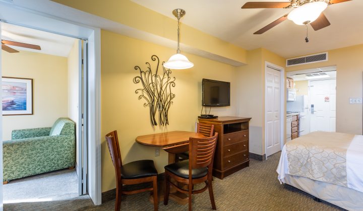 A wide view of a hotel room with a bed, table, chairs, TV, and kitchenette.