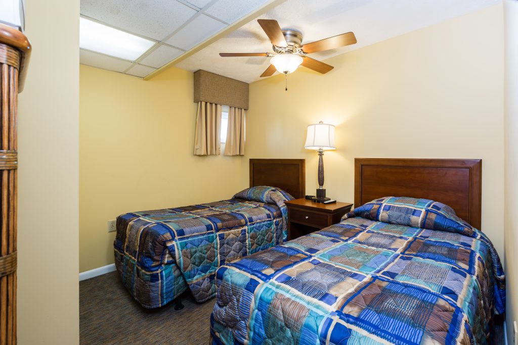 A hotel room with two twin beds, wooden headboards, and patchwork quilts, with a ceiling fan and lamp on a nightstand.