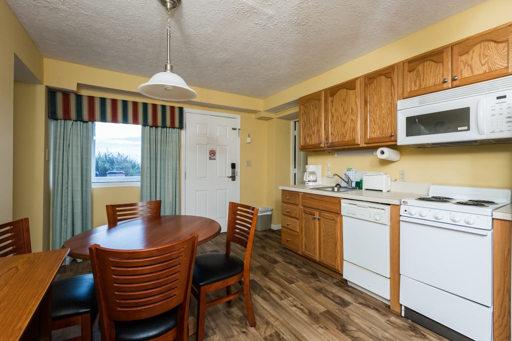 A kitchenette with white appliances and wood cabinets, a dining table with four chairs, and a window with a view.
