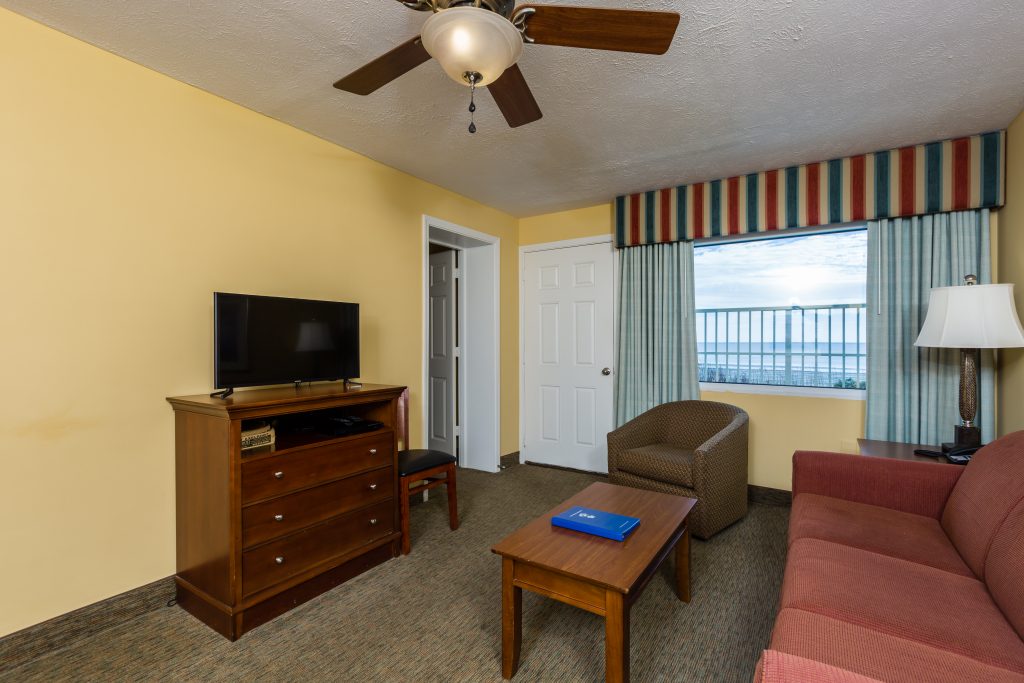 A wide view of a hotel room's living area with a sofa, armchair, coffee table, TV, and window.